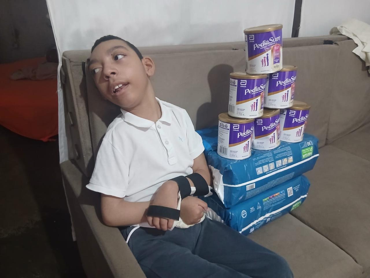 Criança atendida pelo ICCA sentada no sofá, ao lado de latas de PediaSure e pacotes de fraldas doados